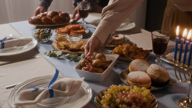 Close up shot of hands of couple placing traditional Jewish dishes on festive dinner table with lit menorah while preparing for Hanukkah celebration at home