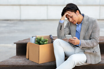 A jobless Asian man in a gray blazer and white pants sits on a wooden bench. He has his belongings in a cardboard box beside him. He looks down at his phone with a worried expression.