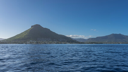 Picturesque mountains against the blue sky and clouds. City houses, green vegetation on the ocean shore. Ripples on the surface of the water. Mauritius. View from the sea