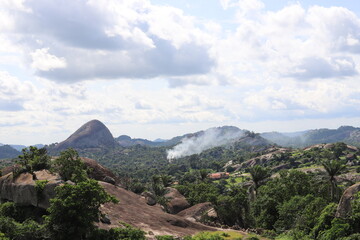 A picturesque view of rolling rocky hills covered in lush greenery under a partly cloudy sky. Smoke...