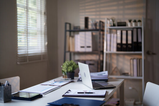 An organized office desk with a laptop, documents, and a small potted plant, set against a background of shelves and window blinds