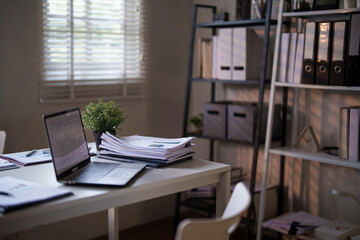 An organized office desk with a laptop, documents, and a small potted plant, set against a background of shelves and window blinds