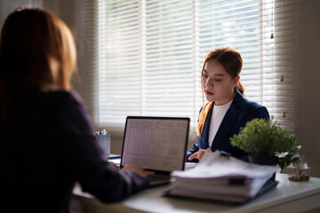 Two businesswomen in suits work at a desk, both focused on their laptops and documents. They are in a professional office setting