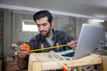 young carpenter caucasian man using measuring tape looking wood size with laptop at workspace. craftsman profession in wood factory.