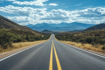Naklejka premium A long, straight road leads through a desert landscape towards a mountain range under a blue sky with clouds.