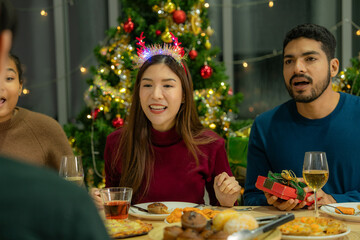 Group of Asian friends enjoying Christmas party for new year celebrate. Friends having fun together at home. men and women opening box gift and present together at home with Christmas tree.