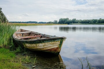 Obraz premium A weathered wooden boat rests on the grassy shore of a tranquil lake, under a partly cloudy sky.