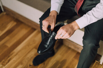 A man is putting on his shoes. He is wearing a black dress shoe and a red tie