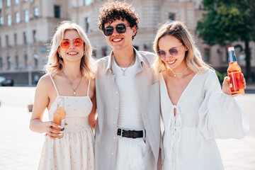 Man and two young beautiful smiling hipster female. Carefree women posing outdoors. Positive models holding and drinking fresh cocktail drink in glass bottle. In the street