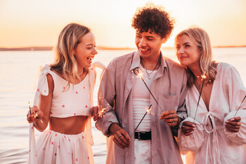 Young three smiling friends having fun during night party at the seaside. Man and two cute female with bengal sparkler lights in their hands. Young women partying on the beach with fireworks at sunset