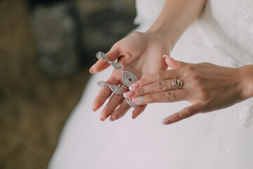 A woman is holding a pair of earrings in her hand. The earrings are silver and have a diamond design. The woman is wearing a white dress and she is getting ready for a special occasion