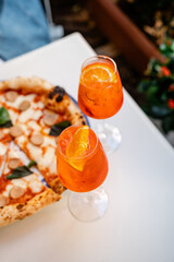 Elegant dining experience featuring two refreshing Aperol Spritz cocktails served in wine glasses, glistening with condensation on a warm day. The drinks sit beside a delicious, wood-fired pizza