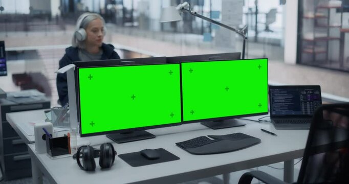 Modern Glass Technology Office Setup with Dual Monitors with Green Screen Mock Up Display, Laptop, Keyboard and Headphones on a Desk. Focused Woman Working in the Background