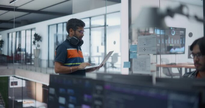 Male Team of Developers, Including a Standing Young Indian Man with a Laptop Computer, Collaborate on a Project in a Modern Glass Office. Colleagues Creating Digital Solutions in a Creative Space