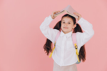 Smiling elementary school student holding a book over her head, wearing a backpack and white shirt, against a pink background