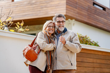 Fototapeta premium Senior couple embracing and smiling in front of a modern house on a sunny day, showcasing warmth, love, and happiness in a relaxed outdoor setting.