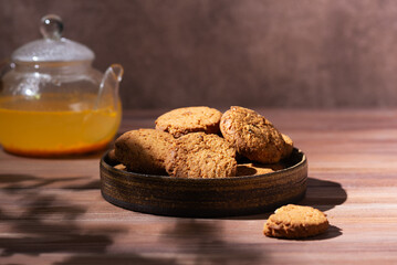 Oatmeal cookies in a plate with sea buckthorn tea in a teapot on a wooden table. Hard light.