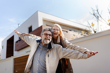 A joyful senior couple poses playfully in front of a modern house, arms outstretched like they’re...