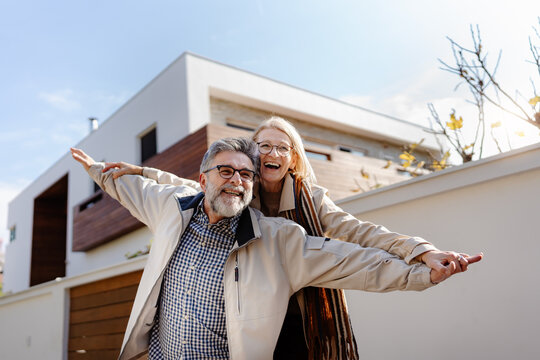A joyful senior couple poses playfully in front of a modern house, arms outstretched like they’re flying, radiating happiness and energy.