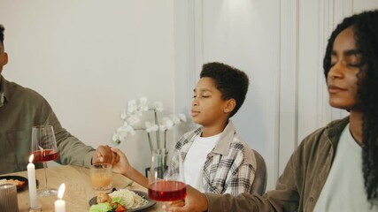 An African American family holds hands and closes their eyes as the father leads a heartfelt prayer of gratitude before their meal, sharing a moment of faith and unity.