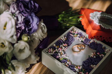 A white and purple bouquet of flowers sits on a table next to a small box with two rings inside. The flowers and rings give off a romantic and elegant vibe