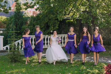 Fototapeta premium A bride and her bridesmaids are walking down a path in a park. The bride is wearing a white dress and the bridesmaids are wearing purple dresses. The bride is holding a bouquet