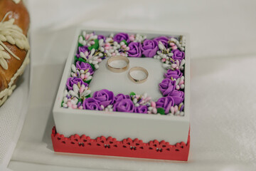 A white box with a purple flower design and two gold rings inside. The box is placed on a table