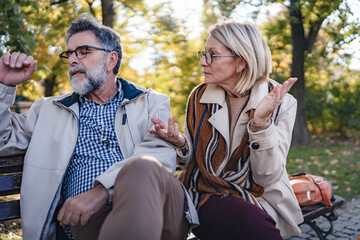 Senior woman appearing to criticize her husband while he sits thoughtfully on a park bench during...
