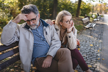 Senior couple sitting apart on a park bench in an autumn setting, looking upset and distant,...