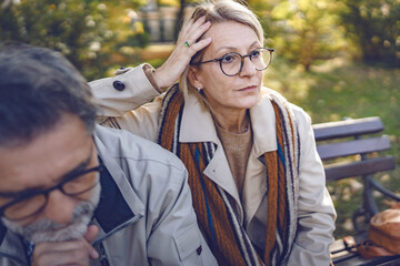 Senior couple sitting apart on a park bench during autumn, looking distant and upset, reflecting...
