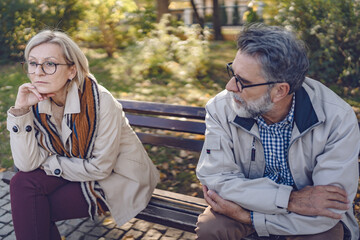 Senior couple sitting apart on a park bench in an autumn setting, looking upset and distant,...
