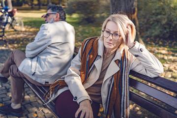 Senior couple sitting back-to-back on a park bench, looking upset and distant, depicting...