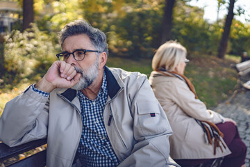 Senior couple sitting back-to-back on a park bench, looking upset and distant, depicting disagreement, relationship challenges, or emotional disconnect in an autumn park setting.