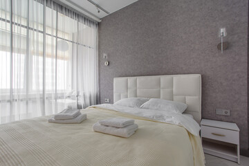 A well-appointed small modern bedroom with a cozy design, featuring a queen-sized bed, bedside tables. A large window with curtains allowing soft daylight.