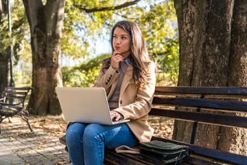 Young businesswoman sitting alone in the park on the bench and working with laptop, making a notes. Business, education, lifestyle concept. Professional occupation female work on computer remotely