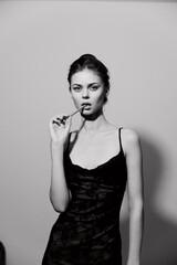 Elegant model in a black slip dress with a seductive expression, holding a pencil and posing against a minimalist light background. Fashion and beauty concept.