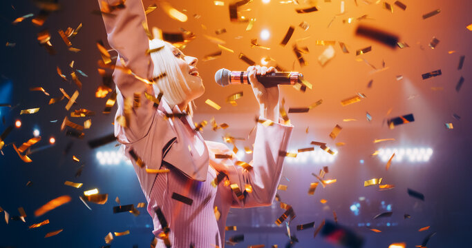Young Female Pop Singer in a Stylish Pink Outfit Performing a Concert on a Festival Stage Surrounded by Falling Confetti. Her Charisma and Lyrics Electrify the Music Fans in Crowded Arena