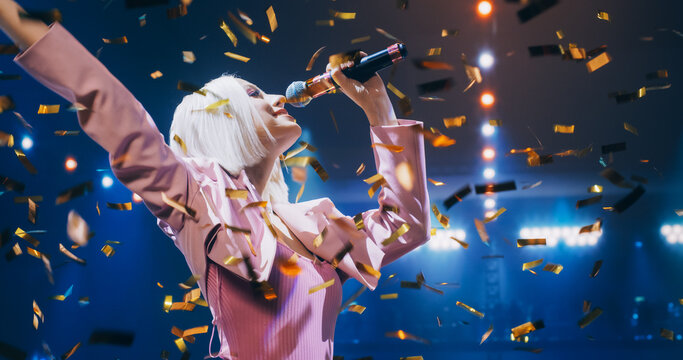 Young Female Pop Singer in a Stylish Pink Outfit Performing a Concert on a Festival Stage Surrounded by Falling Confetti. Her Charisma and Lyrics Electrify the Music Fans in Crowded Arena