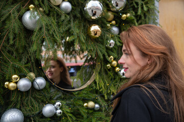 Portrait of beautiful smiling redhead adult caucasian woman in black coat standing by green Christmas tree city street building decorations and looking in reflection on hanging mirror. Soft focus.