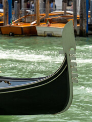 Obraz premium Close-up of a traditional Venetian gondola prow with wooden boats docked in the background on a sunny day in Venice, Italy. Captures the charm of the city's iconic waterways.