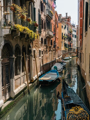 Quaint Venetian canal lined with colorful buildings, balconies with flowers, and moored boats, including a decorative gondola. A classic scene capturing Venice's timeless beauty and charm.