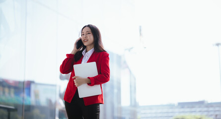 Beautiful Asian businesswoman walking confidently on a modern city street, smiling while using a smartphone. Success, urban lifestyle and modern communication.