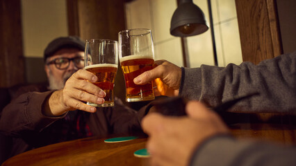 Two elderly men, friends clink glasses of beer in cozy bar, enjoying relaxing evening. Focus on hands and lager beer glasses. Concept of elderly people lifestyle, drinks, retro fashion