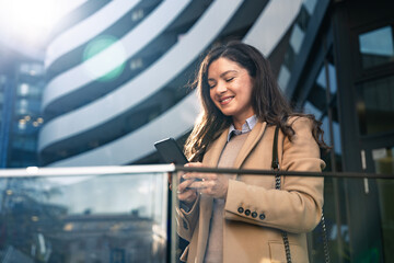 Young adult smiling professional occupation business woman wearing suit holding smartphone while standing in urban city environment. Businessperson female with phone outside of office building.