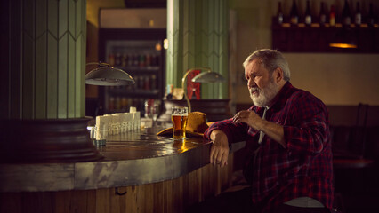 Elderly bearded man in checkered shirt sitting at warm lit bar, drinking beer, talking to bartender and having relaxing time. Concept of elderly people lifestyle, drinks, retro fashion