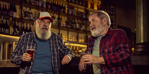 Two elderly men in checkered shirts sitting at modern cozy bar, drinking beer, involved in conversation, and enjoying meeting. Concept of elderly people lifestyle, drinks, retro fashion