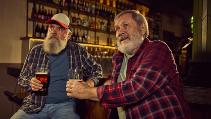 Two elderly men in checkered shirts sitting at modern cozy bar, drinking beer, and looking sport...