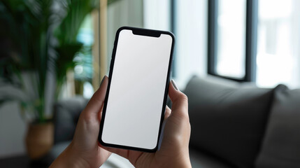 person holding blank smartphone in modern living room setting, showcasing minimalist design and natural light. background features greenery and soft furnishings