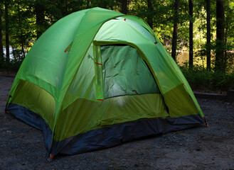 Lighted camping tent in a forest with the sun shining on a lake behind the trees. 