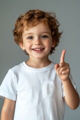 Smiling young boy in a white shirt, pointing up with an enthusiastic expression against a neutral background.